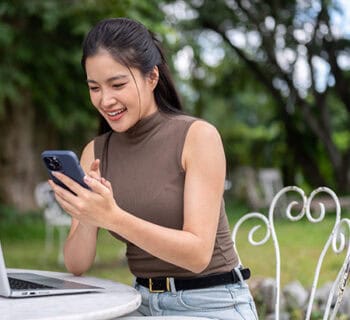 A woman sitting at a park picnic table with her laptop open and phone in her hand signs up for a Lafayette Federal Credit Union online mobile banking account after switching her direct deposit.