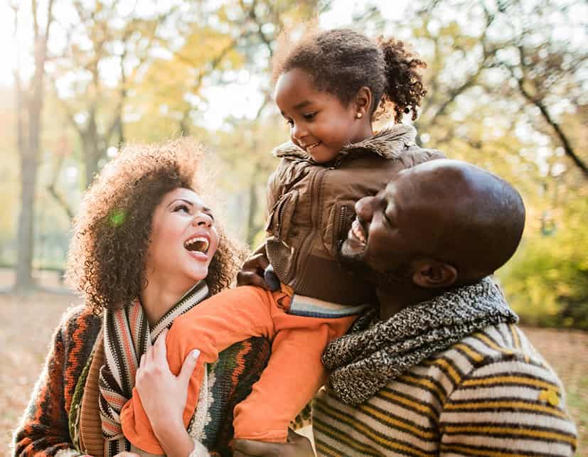 A mother and father hold up their young daughter in on their shoulders in a fall or winter forest.