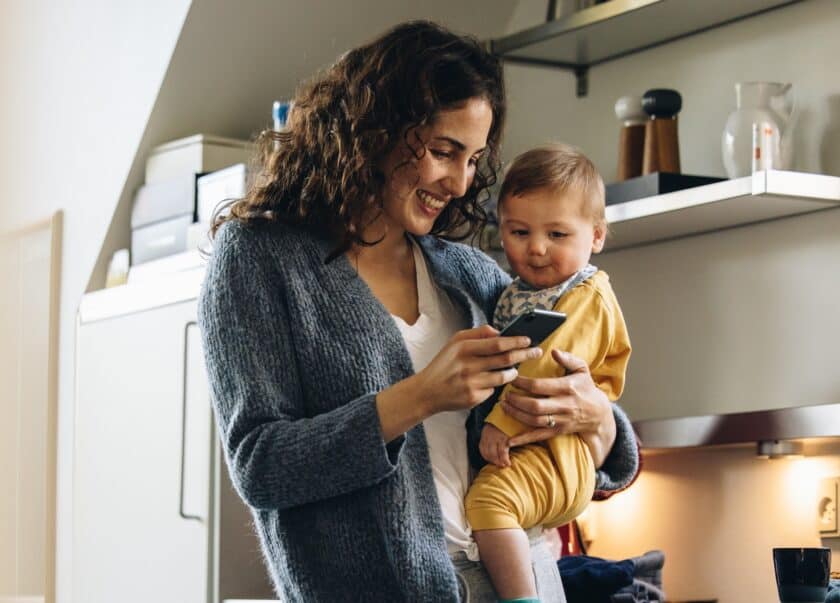 Smiling woman with baby texting on phone at home