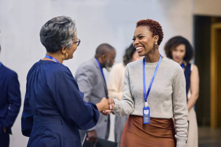 Two women in business attire shaking hands and smiling at a conference. One woman wears a blue dress, the other a gray sweater and brown skirt. Name tags are visible. Background shows other attendees.