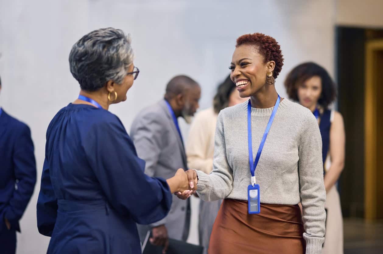 Two women in business attire shaking hands and smiling at a conference. One woman wears a blue dress, the other a gray sweater and brown skirt. Name tags are visible. Background shows other attendees.